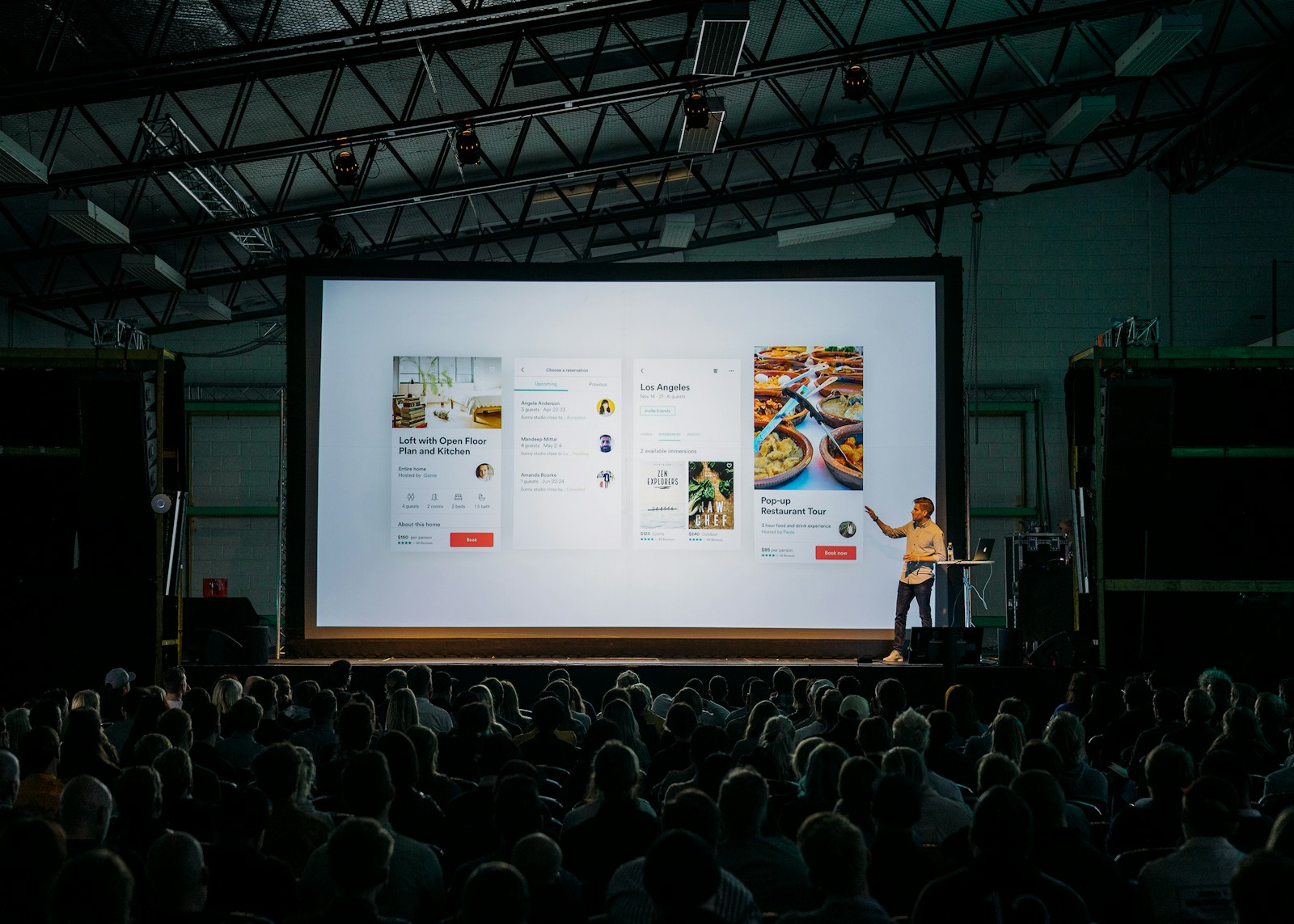 Photo by Teemu Paananen person discussing while standing in front of a large screen in front of people inside dim-lighted room